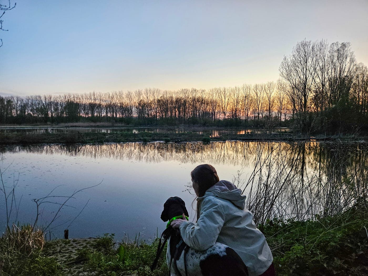 Een vrouw zit met haar hond aan het water bij zonsondergang